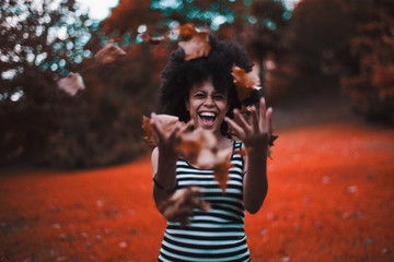 Naklejka premium Curly laughing African-American girl in a striped dress is fooling and throwing up a heap of dry autumn leaves, with forest and meadow in unnatural vivid red colors behind, strong teal bokeh