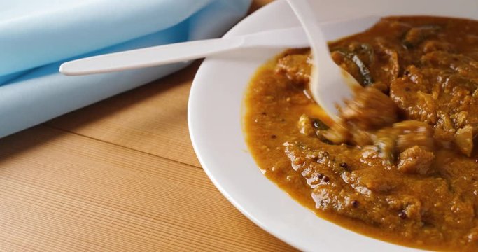 Taking a bite of eggplant curry on a white plate with a plastic fork and knife and a blue napkin to the side atop a wood table top.