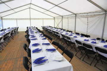 View of rented tent ready for guests. White table cloth, white plates with blue napkins and empty glass. Party celebration concept. 