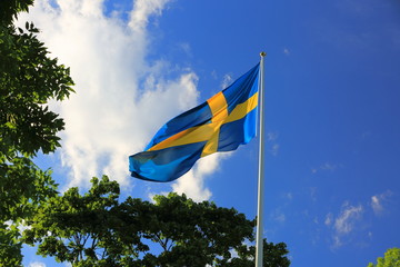 Beautiful view of swedish flag on green trees and blue sky with white clouds background. 
