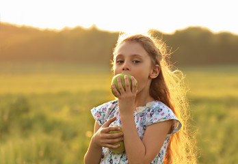 Fun beautiful kid girl with long hair joying and biting green apples on summer bright background. Closeup bright portrait