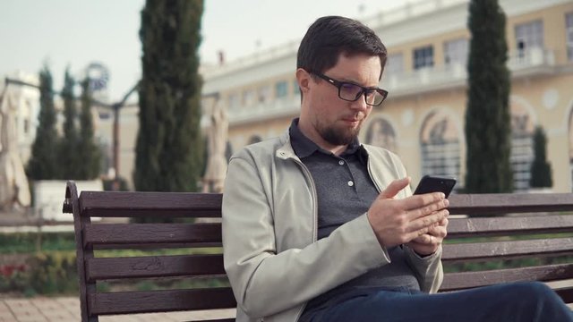 Gay with glasses on face is resting in square, looking on a screen of mobile phone. he is sitting on a bench and texting in daytime