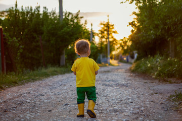 a little boy walks along the country road in rubber yellow boots and a bright T-shirt.