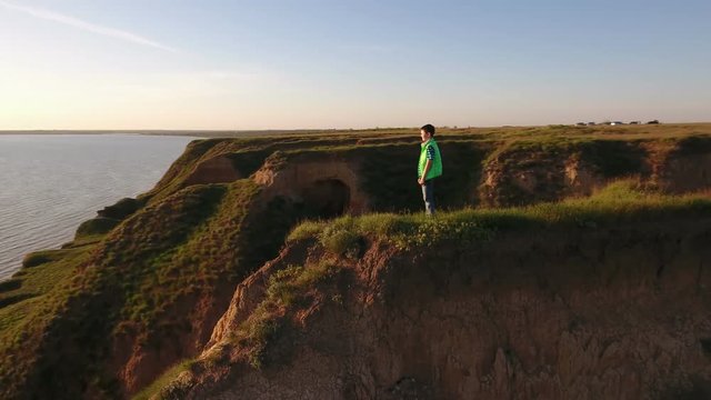Aerial pan of a thoughtful boy standing on a spiky hill with deep gaps nearby at the Black Sea and looking at a glitzy sun path in summer