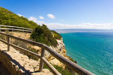 Fototapeta premium Wood railing along walk path of nature park by the sea with Barbate town on the background in Cadiz, Andalusia. Hiking, trek adventure, coastline landscape concept