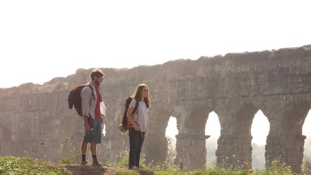 Young lovely couple backpackers tourists reading map pointing directions roman aqueduct arches in parco degli acquedotti park ruins in rome on romantic misty sunrise with guitar and sleeping bag slow