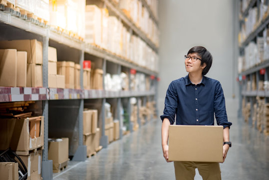 Young Asian man carrying cardboard box between row of shelves in warehouse, shopping warehousing or working pick and packing concepts