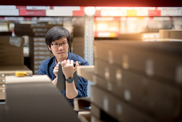 Young Asian man doing stocktaking of product in cardboard box on shelves in warehouse by using digital tablet. physical inventory count concept