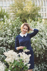 Attractive girl stands by bushes of white lilac and holds flowers in her hands. Evening walk after work. Woman in office style.