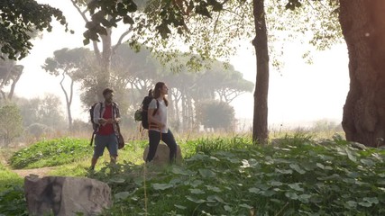 Young happy couple backpackers tourists walking in the nature along dirt road in aqueduct park ruins in rome countryside on misty morning with guitar and sleeping bag slow motion - Powered by Adobe