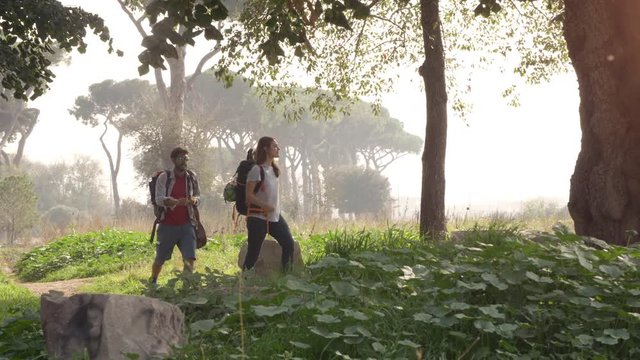 Young happy couple backpackers tourists walking in the nature along dirt road in aqueduct park ruins in rome countryside on misty morning with guitar and sleeping bag slow motion