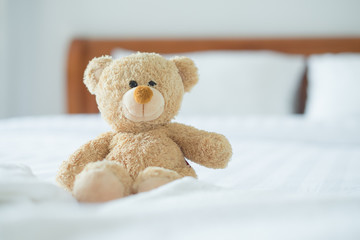 Cute little teddy bear sitting alone on white bed in morning.