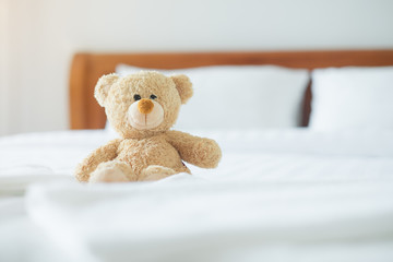 Cute little teddy bear sitting alone on white bed in morning.