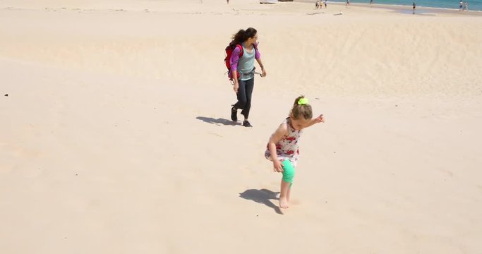 four years old blonde girl with dress with brunette woman mother with backpack walking at beach sand in Cadiz, Andalusia, Spain
