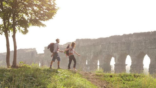Young lovely couple backpackers tourists walking holding hands arrive on hill in front of roman aqueduct arches in parco degli acquedotti park ruins in rome on romantic misty sunrise with guitar and