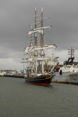 A three-masted modern luxury clipper with historical nineteenth century styling sailing in the North Sea Canal's mouth under the cloudy gray sky, IJmuiden, the Netherlands.