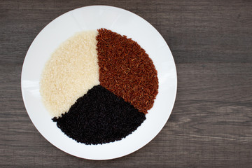 Raw white, black and brown rice on a white plate on a wooden table. Healthy diet food
