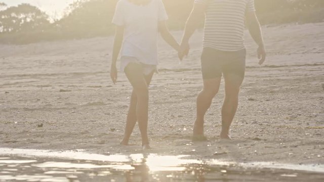 Tilt down of boyfriend and girlfriend holding hands and walking along ocean beach at sunset