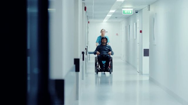Female Nurse Pushing Patient in the Wheelchair Through the Hospital Corridor, They are Going to the Procedures