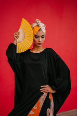 Obraz premium Portrait of a fashionable Muslim woman model in a head scarf hijab against a red background in a studio. She is young, attractive and beautiful and is posing with a yellow fan. 