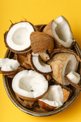 Cracked coconut and coconut pieces arranged in metallic tray on yellow background. Overhead view