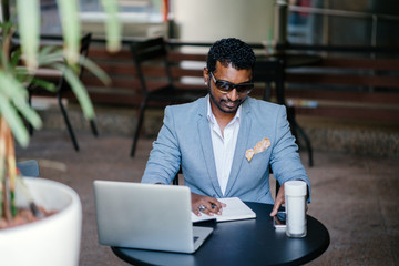 A millenial Indian Asian businessman is writing down ideas in his notebook. He is sitting in a coworking space with his laptop, smartphone, and a flask of coffee nearby.