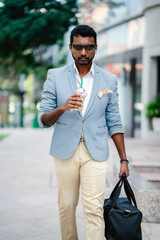 A young and handsome Indian Asian man walks down a street in Asia while drinking a coffee and holding his gym bag. He is dressed smartly and fashionably in a white shirt, blue jacket and khakis. 