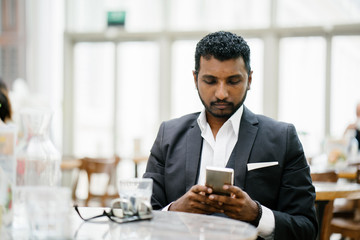 An Indian Asian businessman takes a break from work as he sits in a cafe fiddling through his smartphone. He is well-dressed in a dark suit.