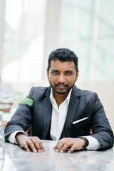 Portrait of a young Indian Asian businessman looking intense and serious at the camera. He is wearing a gray suit and is seated at a table in their office during the day.