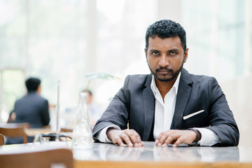 Portrait of a young Indian Asian businessman looking intense and serious at the camera. He is wearing a gray suit and is seated at a table in their office during the day.