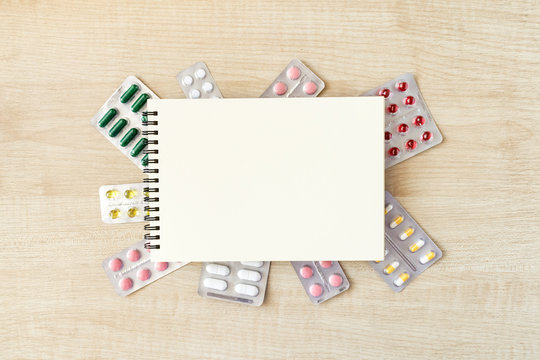 Paper notepad mock up in frame of various blisters with multi-colored tablets and capsules on wooden table. Doctor desk background, horizontal medical concept copy space, white mockup