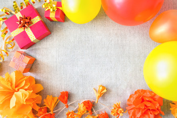 Birthday party background with festive decor, orange, yellow and red ribbons, gift boxes, balloons, garland, pompoms on neutral fabric. Colorful celebration background, copy space, Flat lay, top view.