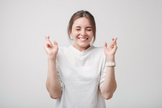 Indoor shot of attractive european girl in white shirt having excited, superstitious look, keeping fingers crossed.