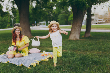 Fototapeta premium Little cute child baby girl with basket play and have fun, rest with woman in yellow clothes on green grass in park. Mother, little kid daughter. Mother's Day, love family, parenthood, childhood.