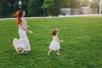 Naklejka na ściany i meble Laughing woman in light dress running catching up with little cute child baby girl on green grass lawn in park. Mother, little kid daughter. Mother's Day, love family, parenthood, childhood concept.
