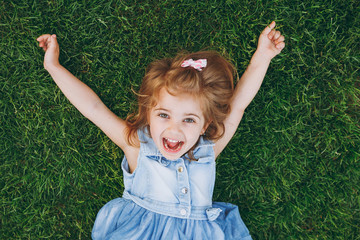 Laughing little pretty child baby girl in light dress lie on green grass lawn, spreading hands in...