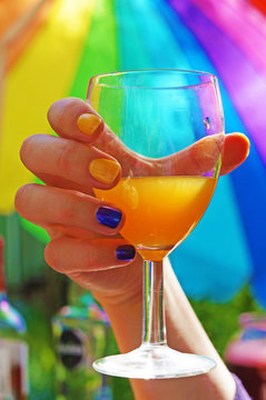 Hand with multi-colored nails holding a glass with a orange cocktail on the background of an rainbow umbrella on a bright sunny day.