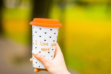 Ceramic cup for coffee on a blurred background of nature. Girl in a hand holds a cup of coffee. concept of coffee take-away. With the text on the cup l love you more. Solar bright saturated image.