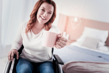 I love coffee. Delighted crippled woman sitting in a wheel chair and holding a cup of coffee