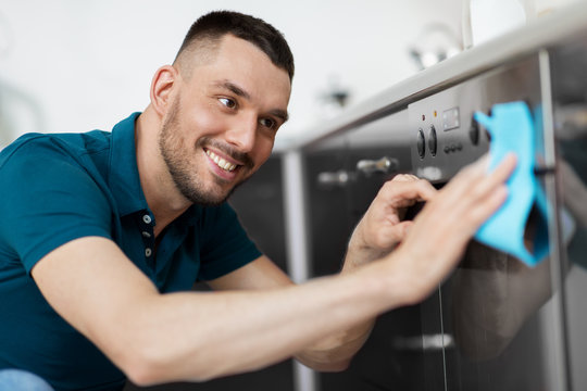 household and people concept - happy smiling man wiping table with cloth cleaning oven door at home kitchen