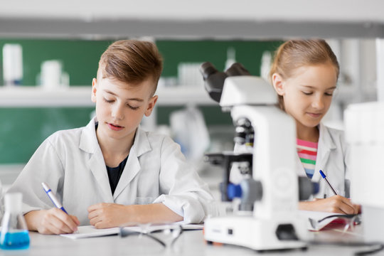 education, science and children concept - kids studying chemistry at school laboratory and writing to workbooks