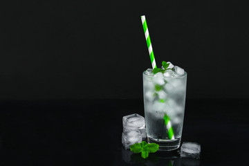 Ice water with ice and a leaf of mint and a straw in a glass on a black background.