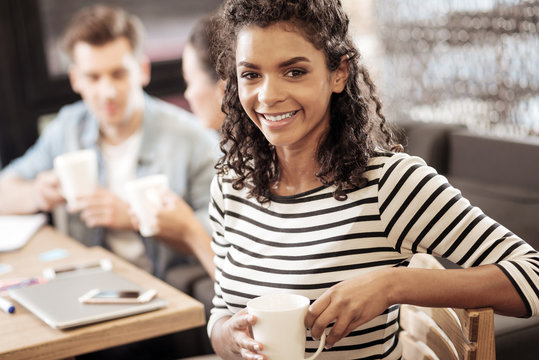 Tasty drink. Attractive cheerful woman holding a cup of tea while having a lunch break with her colleagues