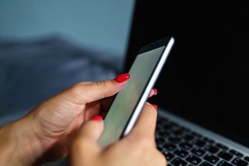 Close-up of a girl with a nice manicure touches the screen of the smartphone on the background of a laptop in the bedroom. Concept of high angle cheating, mobile banking, online payment.