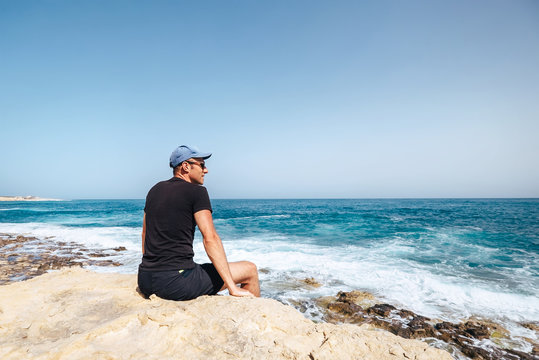 Man sits on rocky cliff near the seaside and looks on waves