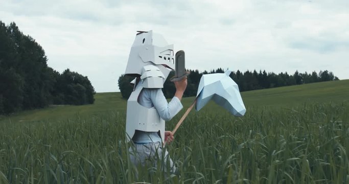 Kid boy wearing cardboard medieval knight armor costume riding stick horse through a grass field in summer. 4K UHD 60 FPS SLO MO