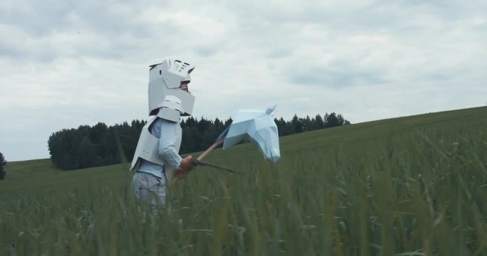 Kid boy wearing cardboard medieval knight armor costume riding stick horse through a grass field in summer. 4K UHD 60 FPS SLO MO