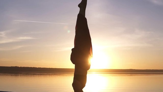 Close up footage of a beautiful girl standing stood on her head in yoga asana. Sun shines and water on the background