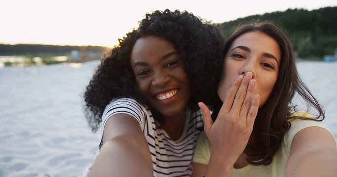 POV of two multiethnical cheerful female friends making nice smiles and showing victory sign with fingers. At the lake. Outdoor