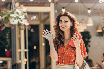portrait of cheerful woman with smartphone in hand waiving to someone on street
